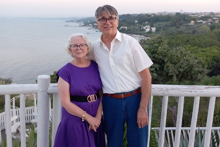 Mario and Sheila pose near a white-railed seaside overlook, with ocean, lush greenery, and coastal homes behind them in soft evening light.